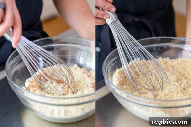 A whisk is actively blending the dry ingredients for paleo pumpkin muffins in a clear, large mixing bowl, showing the almond flour, spices, and leavening agents being incorporated.