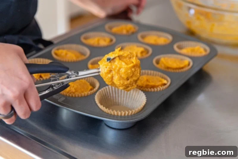 Someone is using a disher (cookie scoop) to evenly scoop the paleo almond flour muffin batter into the parchment-lined muffin tin, ensuring consistent portion sizes.