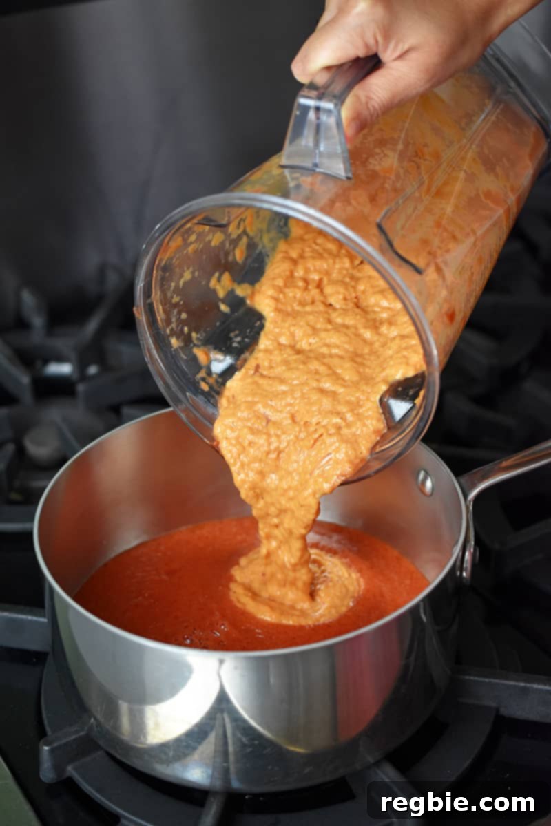 The blended mixture of leeks, garlic, and tomato purée being poured from a blender into a saucepan, combining with more puréed tomatoes.