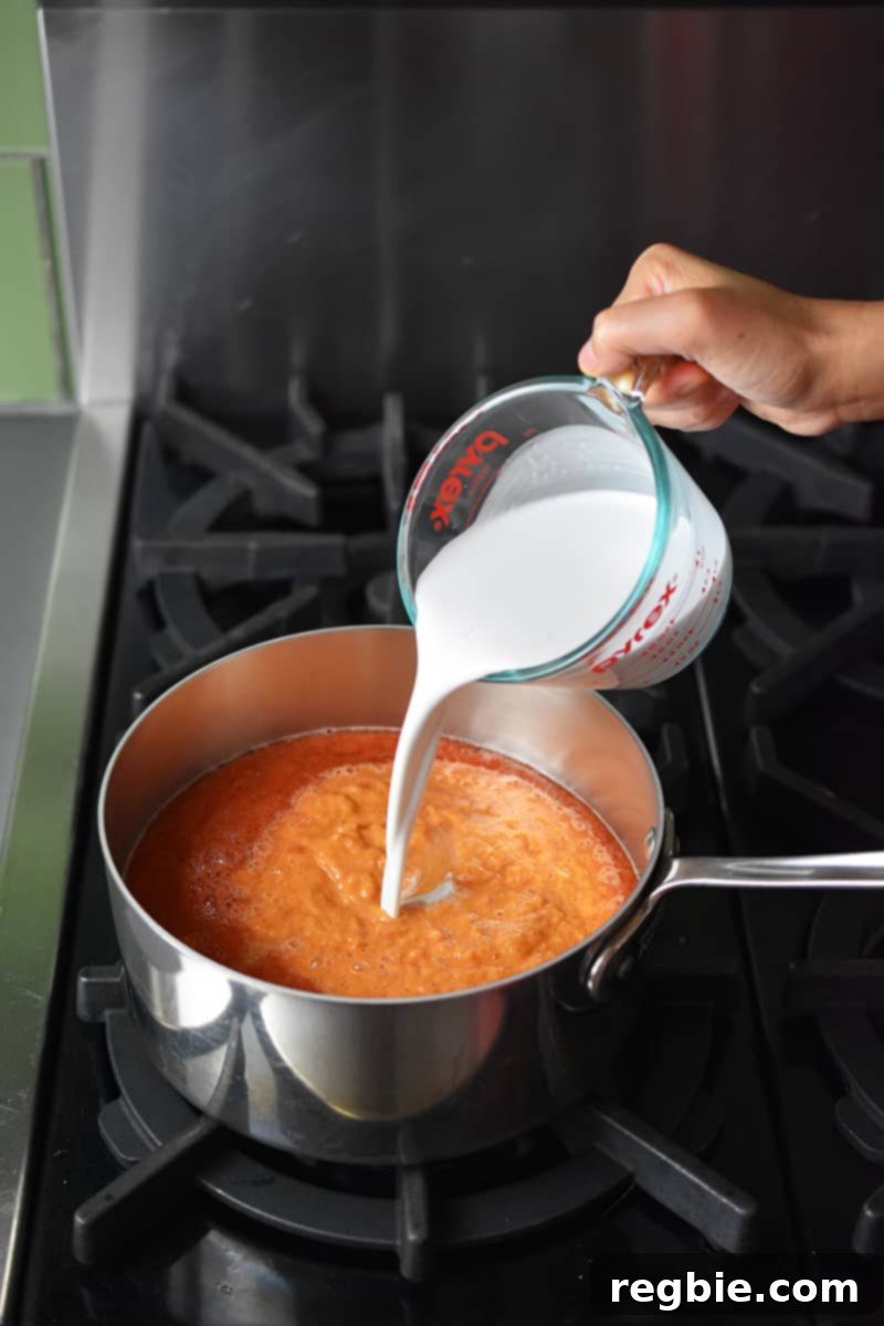 Pouring rich, full-fat coconut milk into a saucepan filled with creamy tomato soup base.