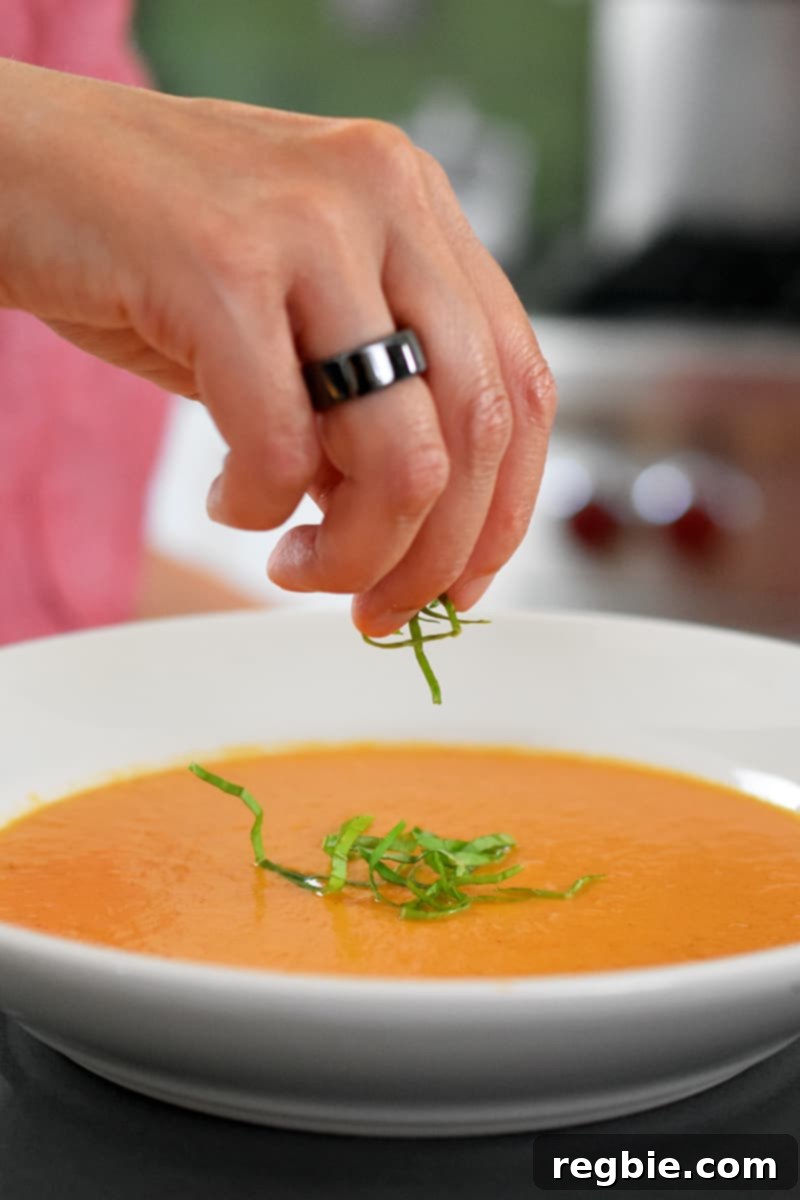 A hand sprinkling finely minced fresh basil over a freshly poured bowl of creamy dairy-free tomato soup.