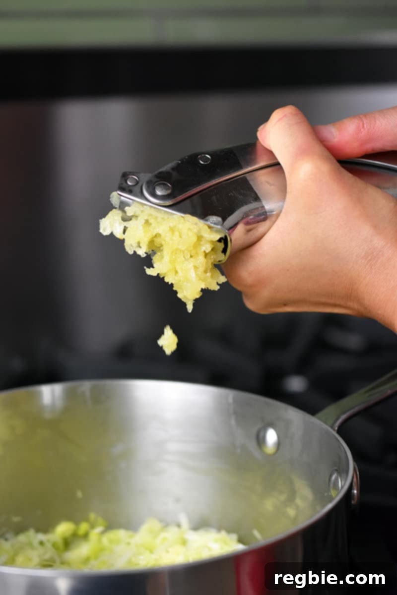 Adding minced garlic from a press to a saucepan filled with softened leeks.
