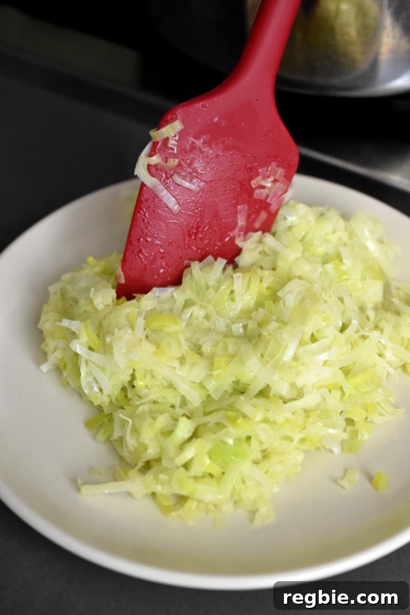 A red spatula transferring cooked leeks and garlic from a saucepan to a clean plate.