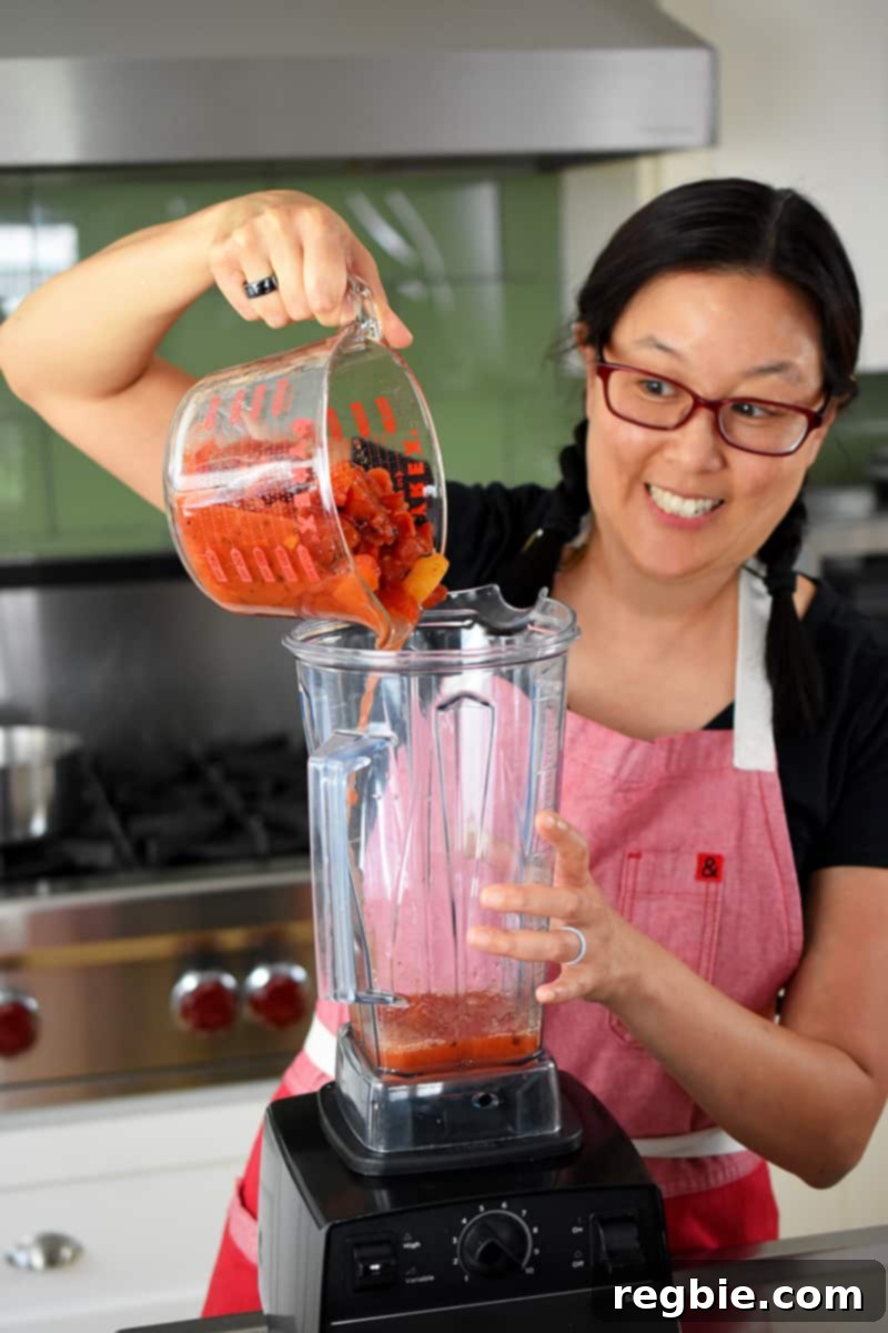 An individual pouring diced canned fire-roasted tomatoes into a Vitamix blender for puréeing.
