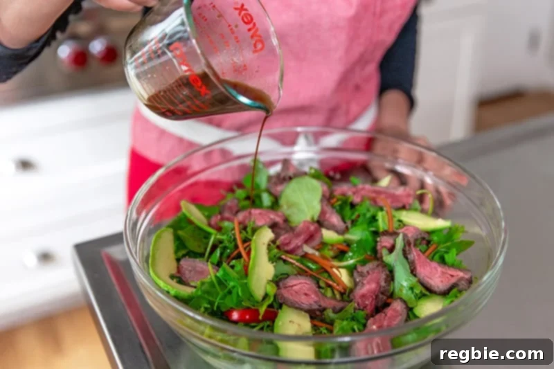 Pouring salad dressing on the Asian Steak salad that has sliced steak, avocados, and vegetables in a clear bowl.