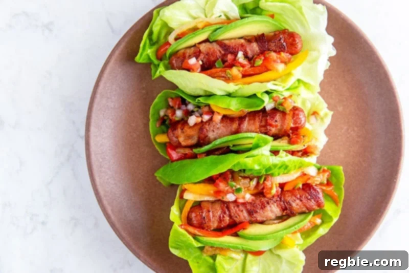 A brown plate with three lettuce-wrapped bacon-wrapped Sonoran hot dogs topped with salsa, sliced avocado, peppers, and onions.