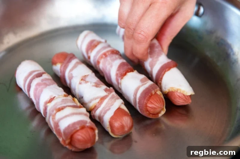 Four bacon-wrapped hot dogs are placed in the middle of a stainless steel skillet to make Sonoran hot dogs.