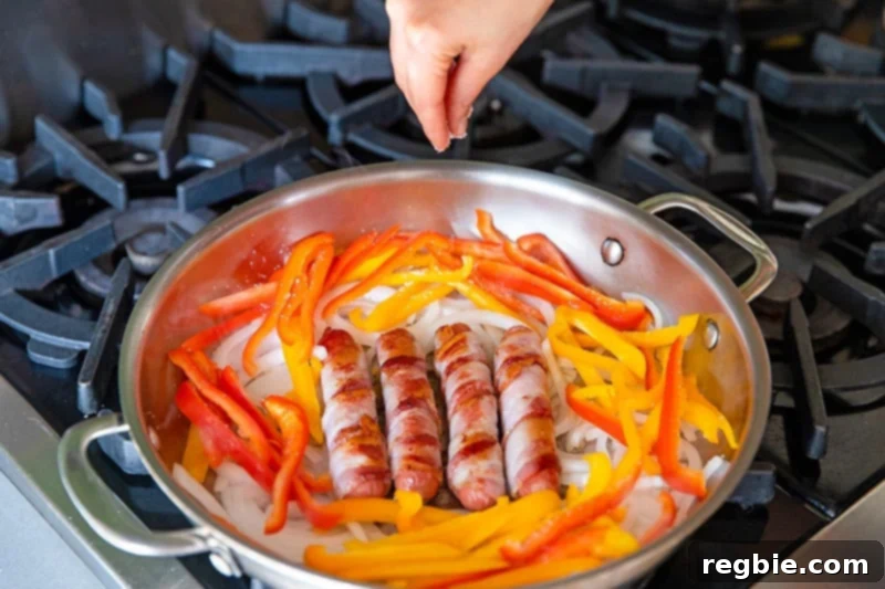 An overhead shot of a skillet with bacon-wrapped Mexican hot dogs surrounded by sliced onions and red and yellow bell peppers. A hand above is sprinkling salt on the vegetables.