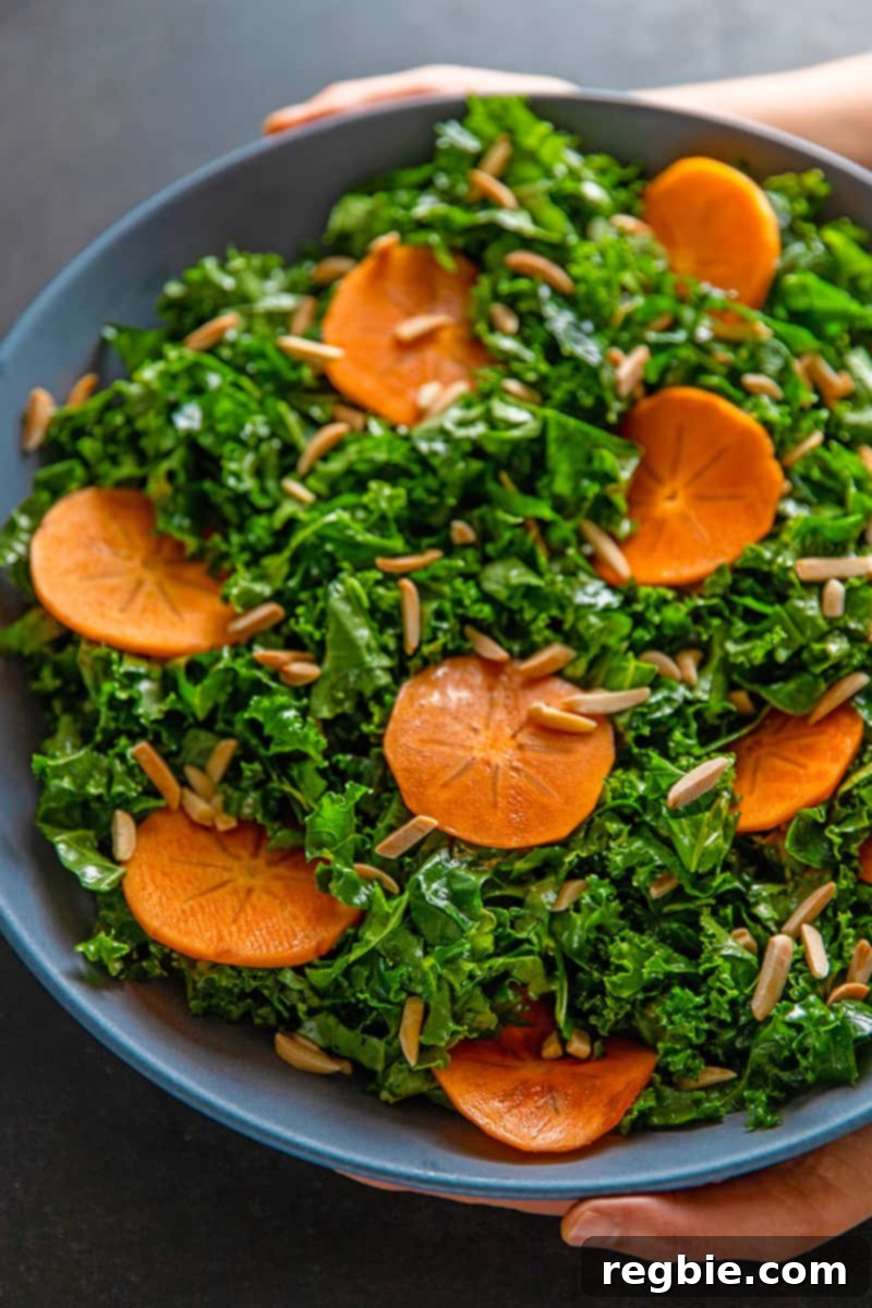 An overhead shot of two hands holding a large blue bowl filled with kale salad and persimmons and toasted almonds, a simple and delicious winter salad.