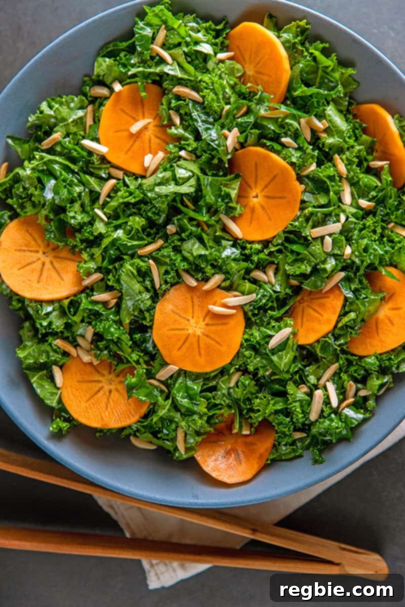 An overhead shot of a large blue bowl filled with Whole30 and vegan kale salad with persimmon next to a pair of wooden tongs.