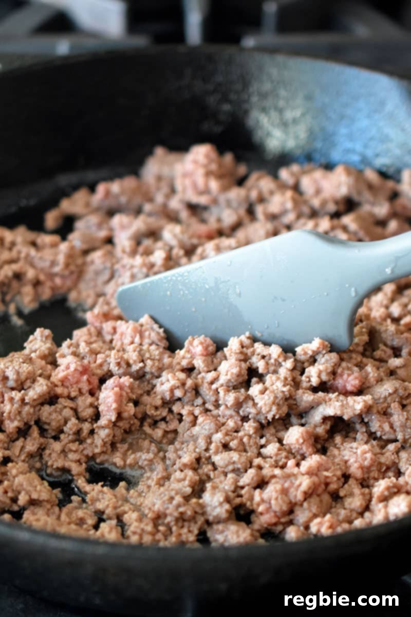 Frying ground beef in a cast iron skillet and stirring it with a gray silicone spatula. The beef is fully cooked and no longer pink.