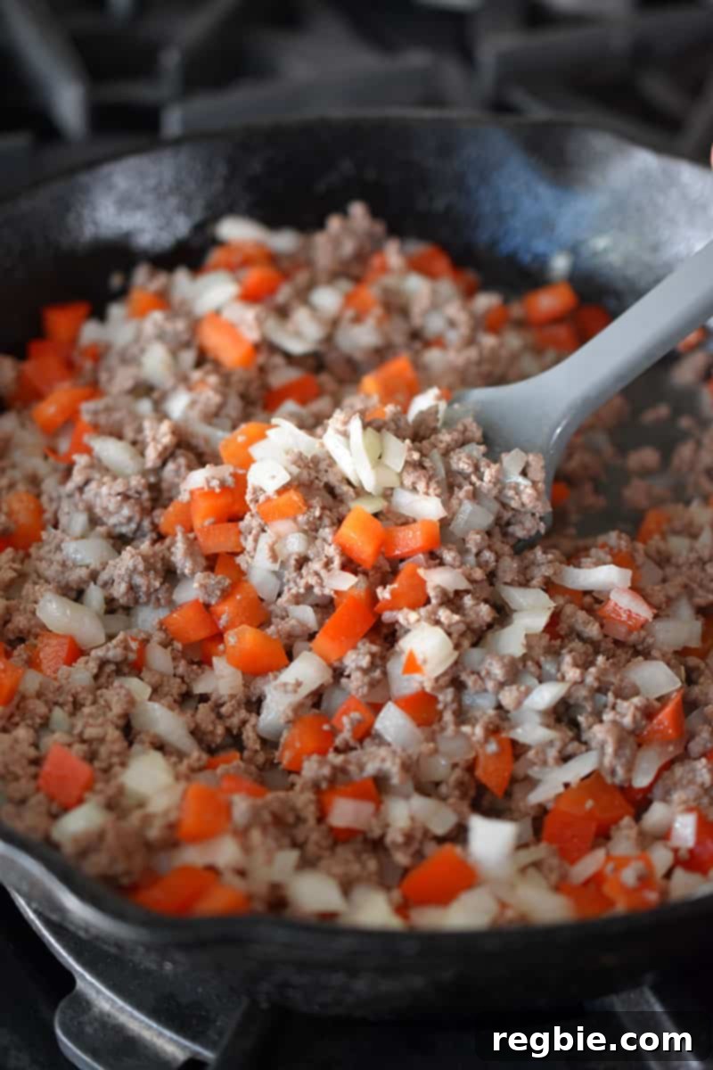 An overhead shot of a cast iron skillet filled with ground beef, diced onions, and red bell peppers.