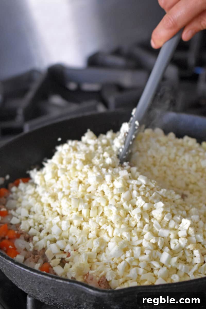 Adding cauliflower rice to a skillet to make the filling for Whole30, paleo, and low carb Tex-Mex Beef and Rice Casserole