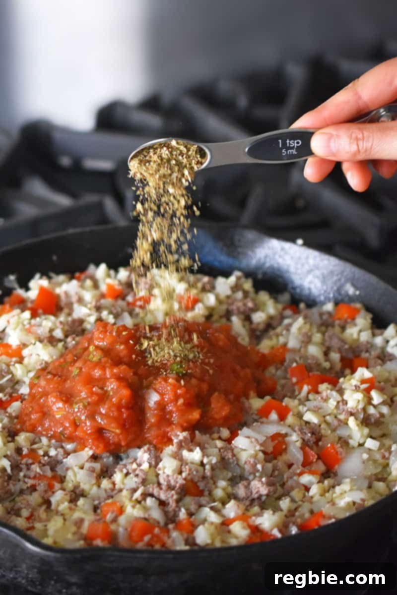 Someone adding a spoonful of oregano to a cast iron skillet filled with ground beef, cauliflower rice, salsa, red peppers and onions.