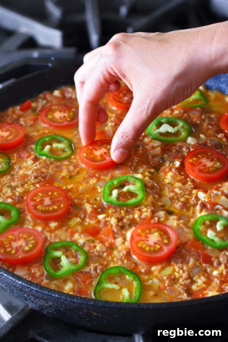 Adding halved cherry tomatoes and sliced jalapeño peppers on top of paleo taco casserole with ground beef and cauliflower rice.