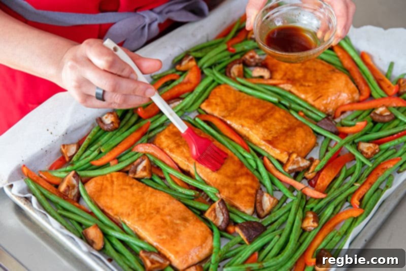 A chef's hand brushing the reserved 'fresh' All-Purpose Stir-Fry Sauce over the finished, glistening Sheet Pan Teriyaki Salmon and roasted vegetables, adding a final burst of flavor and shine.