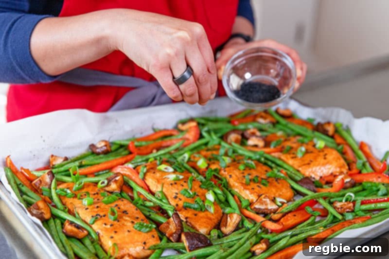 A generous scattering of toasted black sesame seeds being sprinkled over the beautifully cooked Sheet Pan Teriyaki Salmon and roasted vegetables, adding a finishing touch of flavor and texture.