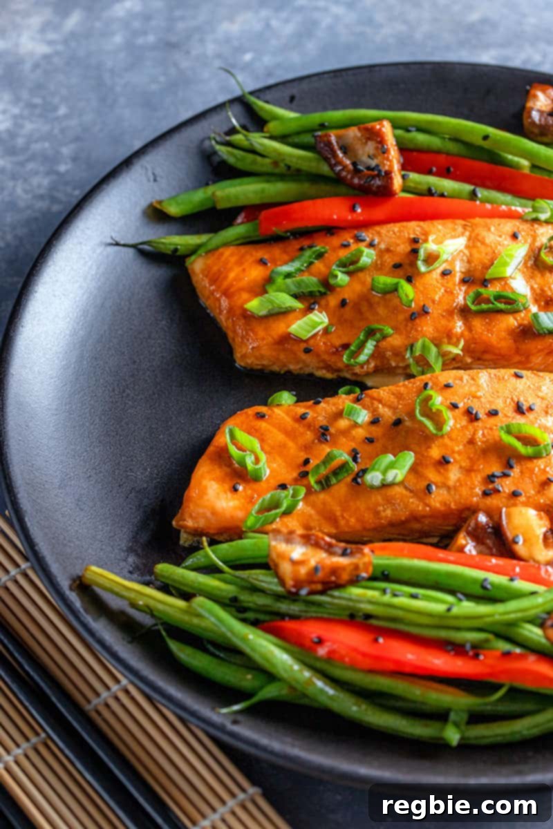 An inviting overhead shot of a sleek black plate showcasing two perfectly cooked Whole30 Teriyaki salmon fillets, artfully arranged with a generous portion of roasted green beans, red bell peppers, and shiitake mushrooms, ready to be enjoyed.