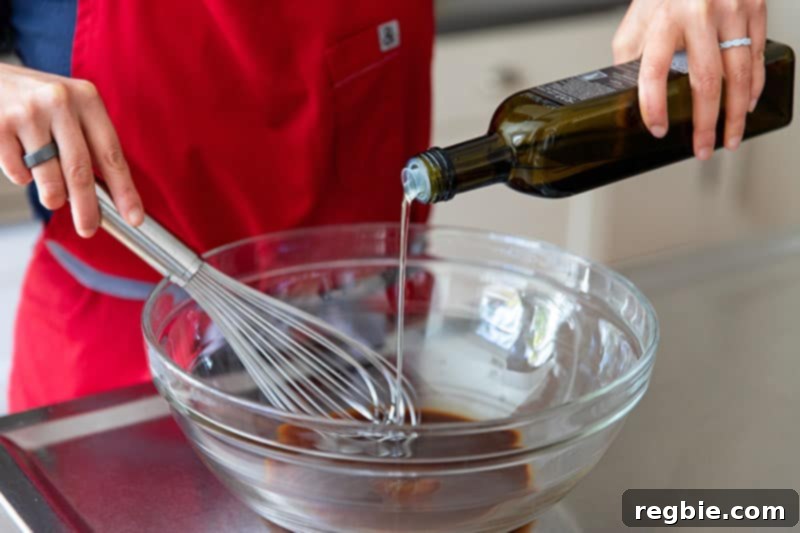 A close-up view of a whisk blending avocado oil and All-Purpose Stir-Fry Sauce in a large bowl, creating the savory sauce for the Whole30 Sheet Pan Teriyaki Salmon's vegetables.
