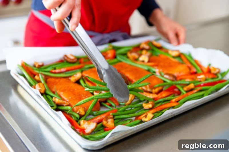 Tossed vegetables, including green beans, red bell pepper, and shiitake mushrooms, meticulously arranged in a single, even layer around the pre-positioned teriyaki salmon on the sheet pan, preparing for oven roasting.