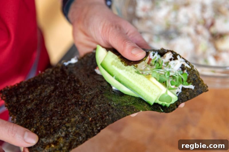 Freshly sliced cucumber, creamy avocado, and delicate sprouts are being carefully layered on top of the seasoned crab salad on a piece of nori, ready for wrapping.