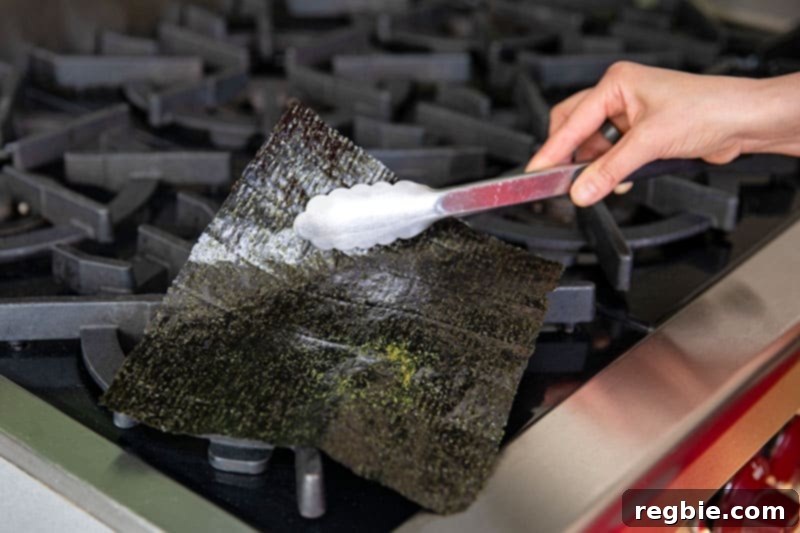 A chef carefully using metal tongs to fan a piece of nori over a low gas burner, toasting it to a vibrant green for homemade California sushi hand rolls.