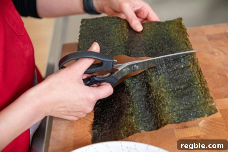 A person skillfully using kitchen shears to cut a freshly toasted sheet of nori in half width-wise, preparing it for assembling Whole30 and keto California hand rolls.