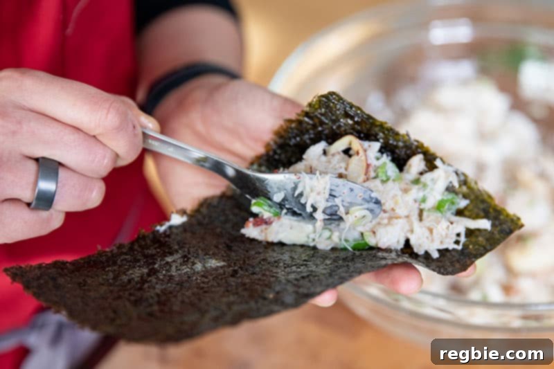 A hand carefully adding the flavorful crab salad filling onto a rectangular piece of toasted nori, preparing to assemble a homemade California hand roll.