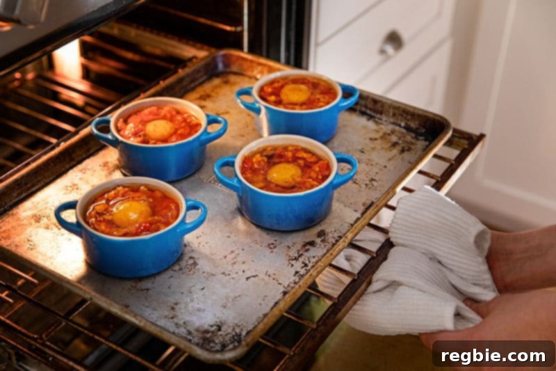 Inferno Eggs 14 A rimmed baking sheet with four blue cocottes filled with eggs in purgatory are being placed into a hot oven.