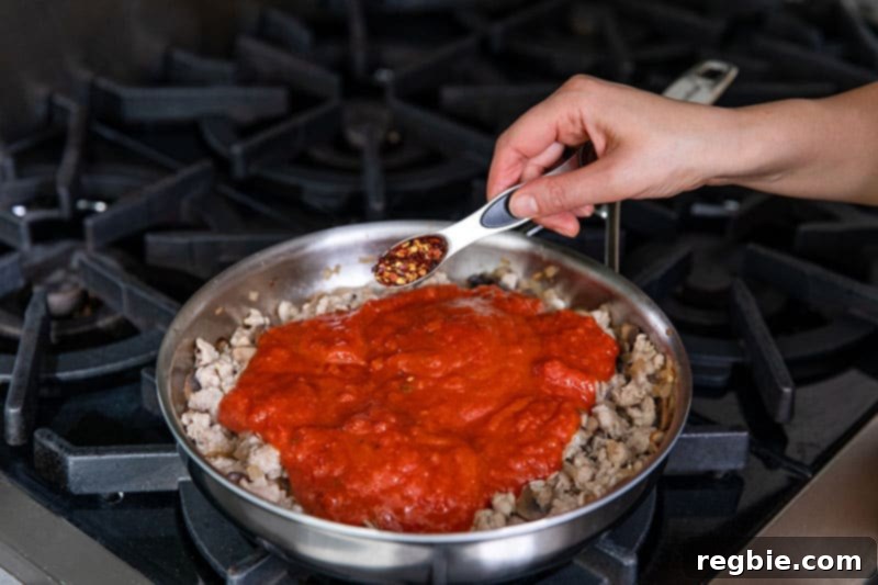Inferno Eggs 9 A hand adding spoonful of crushed red peppers to a stainless steel skillet filled with cooked sausage, mushrooms, and marinara sauce.