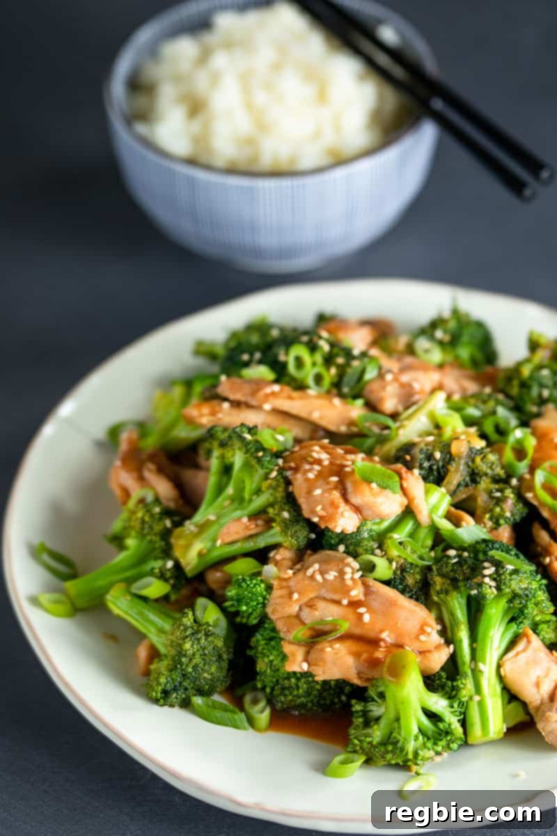 A large white platter generously topped with a vibrant Whole30, paleo, and gluten-free chicken and broccoli stir-fry, garnished with fresh scallions and toasted sesame seeds. In the background, a blue bowl of fluffy cauliflower rice awaits.