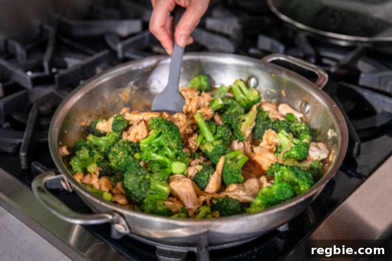 An overhead shot of paleo chicken and broccoli stir-fry cooking in a large skillet, with the sauce simmering and thickening around the ingredients.
