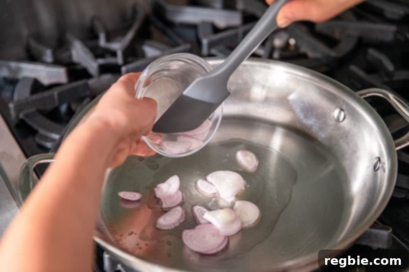 Adding thinly sliced shallots from a clear bowl into a hot stainless steel skillet, beginning to sauté them for the stir-fry base.