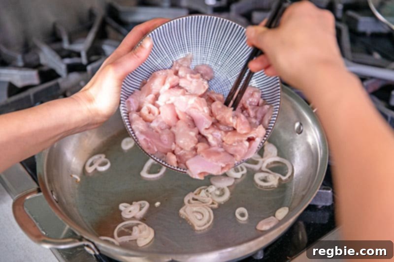 Adding sliced raw chicken thighs to a large stainless steel skillet containing sautéed shallots, ready to be stir-fried.