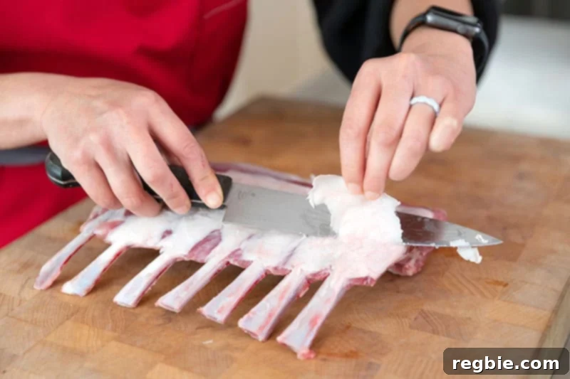 A chef's knife precisely trimming excess fat from the top of a rack of lamb, leaving a thin layer.