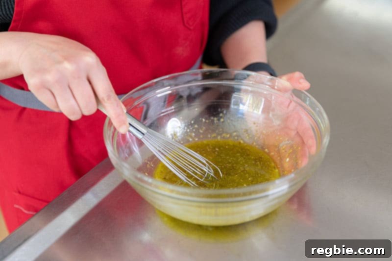 Ingredients for the lamb marinade being whisked together in a bowl: lemon juice, minced garlic, olive oil, dried oregano, and black pepper.