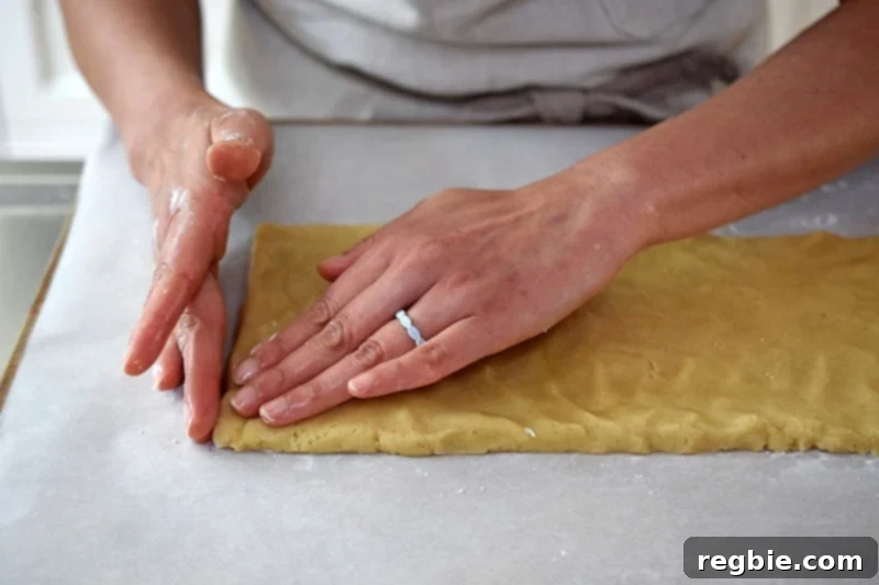 Two hands pressing and shaping paleo cinnamon roll dough into a thin, rectangular form on tapioca-dusted parchment paper.