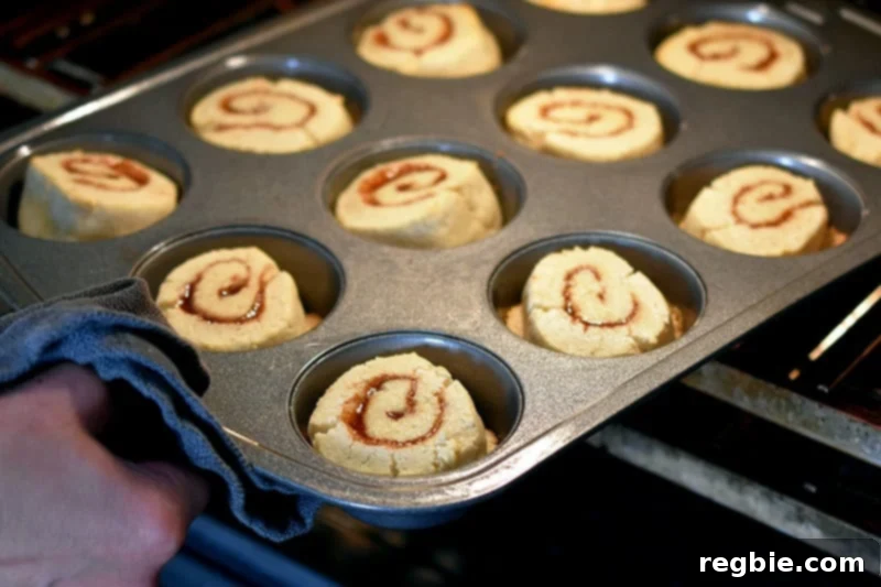 A hand rotating a muffin pan filled with paleo cinnamon rolls halfway through their baking process to ensure even cooking and browning.