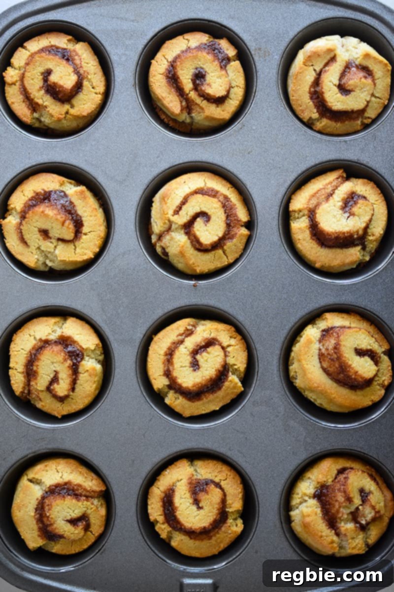 An overhead view of a muffin tin filled with freshly baked, golden-brown paleo cinnamon rolls, indicating they are fully cooked and ready for frosting.