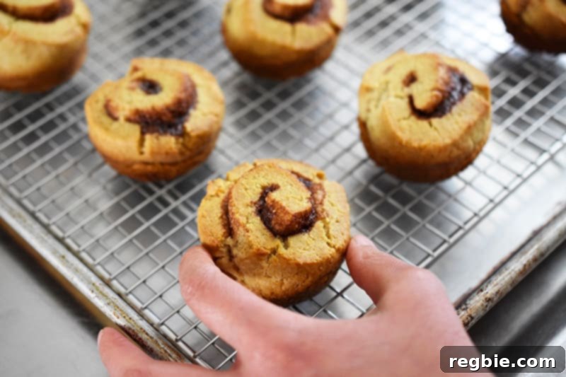 A hand gently placing a warm paleo cinnamon roll onto a wire rack, which rests on a rimmed baking sheet, preparing it for frosting.