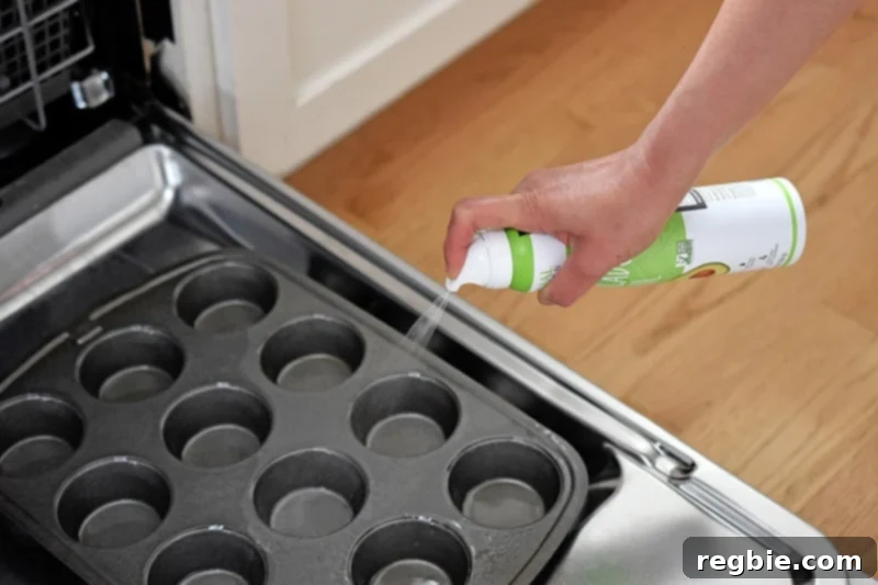 A person spraying a muffin pan with avocado oil while it rests on an open dishwasher door, a smart hack to contain mess.