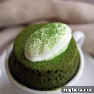 A close-up of a grain-free and gltuen-free matcha mug cake puffed over a white coffee mug and topped with dairy-free coconut whipped cream.