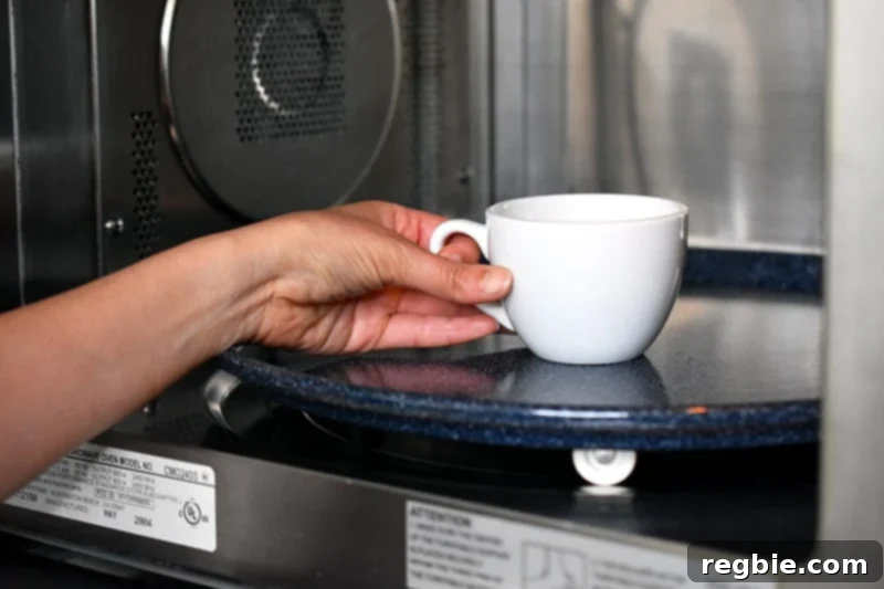 A hand placing a white coffee mug filled with paleo matcha mug cake into an open microwave.