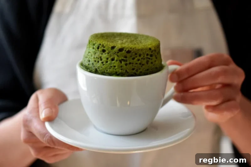 A woman is holding a white coffee mug and saucer. The mug is filled a puffed up green matcha mug cake. 