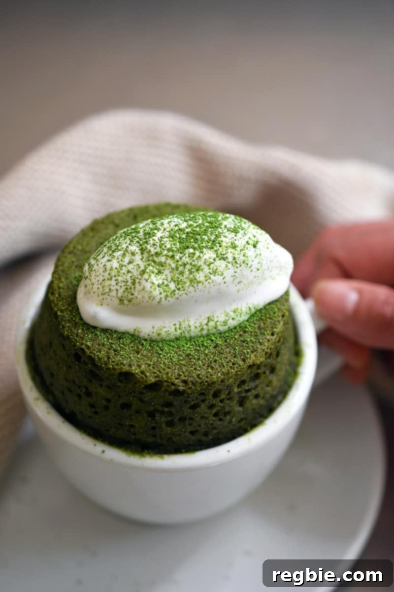 A hand holding the handle of a white mug filled with paleo matcha mug cake and topped with dairy-free whipped coconut cream. 