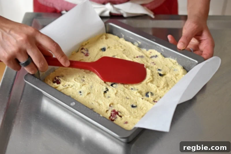 A red silicone spatula is smoothing the top of berry snack cake batter in a parchment lined square metal pan.