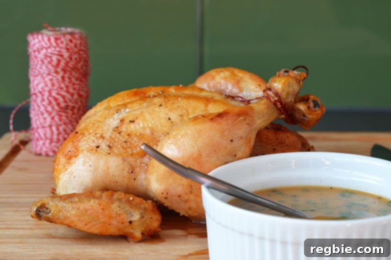 A magnificent oven-roasted whole chicken rests on a wooden cutting board, proudly presented next to a pristine white bowl brimming with its delectable pan sauce.