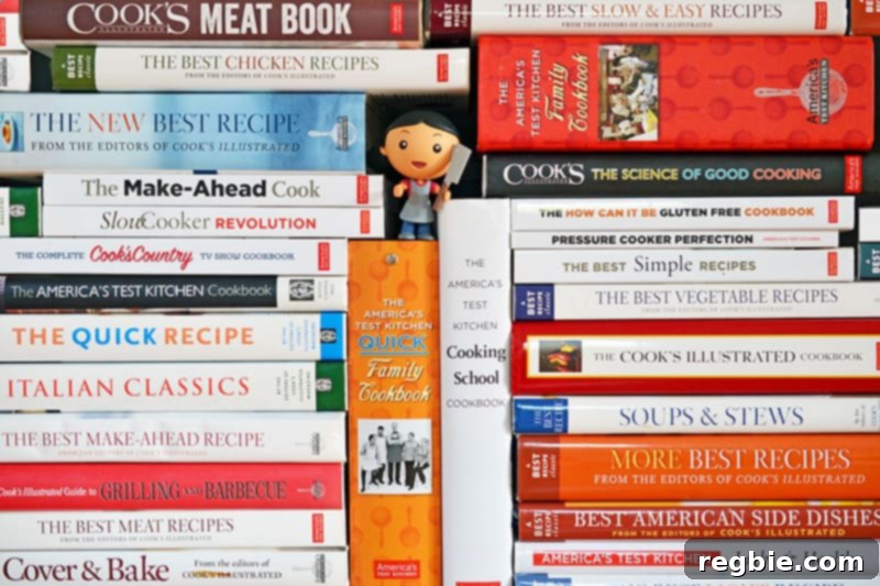 A substantial stack of Cook's Illustrated and America's Test Kitchen cookbooks, showcasing a wide collection of culinary expertise, with a small action figure nestled amongst them.