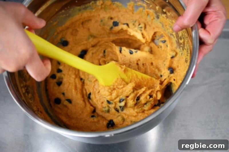 Using a yellow silicone spatula to gently fold dark chocolate chips into the pumpkin muffin batter, ensuring even distribution.