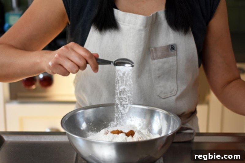 Grain Free Spiced Apple Bundt Cake 8 Someone adding a spoonful of baking soda to a bowl already containing the dry ingredients for the paleo and gluten-free apple Bundt Cake.